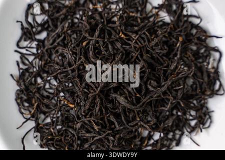 Close-up of dried black tea leaves on a white ceramic plate, showcasing their rich color and texture. Ideal for brewing a healthy, flavorful, and arom Stock Photo