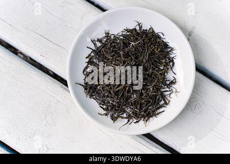 A top-down view of fresh loose leaf tea placed in a white ceramic dish on a rustic wooden table. The tea leaves are black, dry, and intricately curled Stock Photo