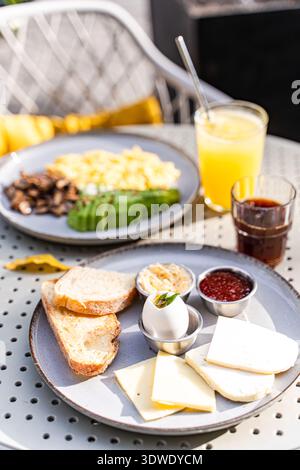 An inviting outdoor brunch setting features sliced cheese, a boiled egg, fresh bread, and accompaniments, complemented by orange juice and coffee for Stock Photo