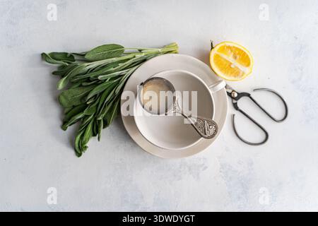 A fresh bundle of sage herb lies beside a white cup and saucer on a textured surface. A half lemon, silver tea strainer, and vintage scissors complete Stock Photo