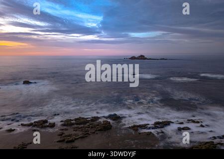Tranquil ocean scene at sunset with a rocky shoreline, gentle waves, and colorful clouds. The serene sky shifts from warm hues to cool blues, embodyin Stock Photo