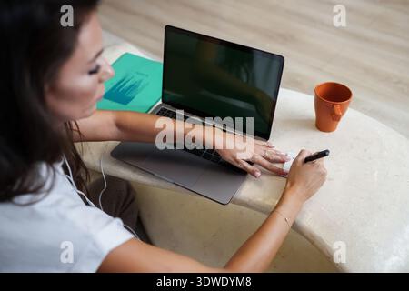 A businesswoman sits at a marble table with a laptop, writing notes on a notepad. An orange mug and a green folder add color to the workspace, creatin Stock Photo