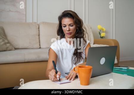 Woman focused on work from home, writing notes while using a laptop at a comfortable living room table, with coffee and phone nearby, creating a produ Stock Photo