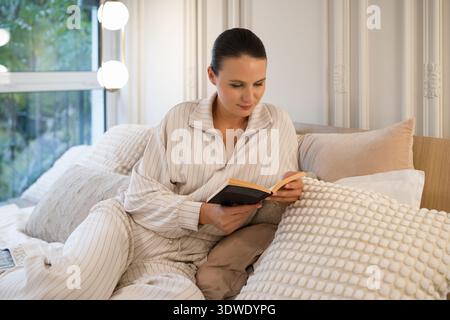 A young woman is comfortably perched on a bed, enjoying a good book in a serene home setting. She is dressed in casual attire, exuding relaxation and Stock Photo