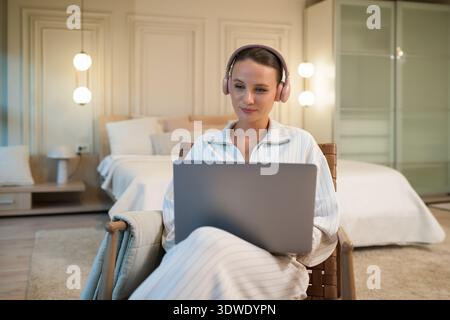 A young woman sits comfortably in her home bedroom, wearing headphones and focused on her laptop screen. Soft lighting and cozy decor create a warm, i Stock Photo
