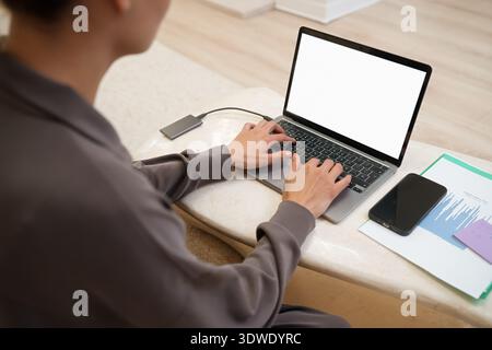 A young woman types on a laptop at home, connected to an external drive. A mobile phone and documents lie nearby on a marble coffee table in a well-li Stock Photo