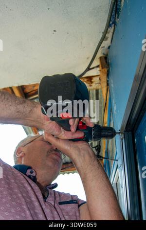 A man with a moustache uses a cordless drill to repair a door frame at home, showcasing DIY skills and home maintenance in a practical setting with na Stock Photo