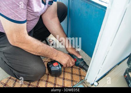 A man kneels on a patterned carpet, focused on using a cordless drill for a home repair project. He carefully drills into the wall to complete the tas Stock Photo