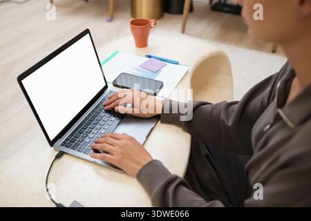 Young woman sitting at a modern beige table in a cozy home, focusing on her laptop. A mobile phone and documents are nearby, suggesting a productive w Stock Photo