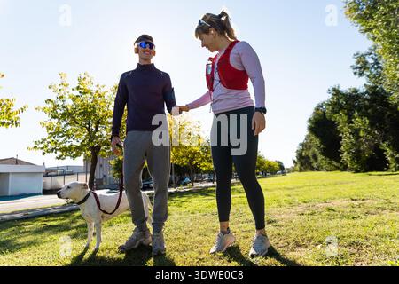 A young man and woman enjoy a sunny day in the park, engaged in outdoor fitness activities. They are dressed in athletic wear and accompanied by a dog Stock Photo