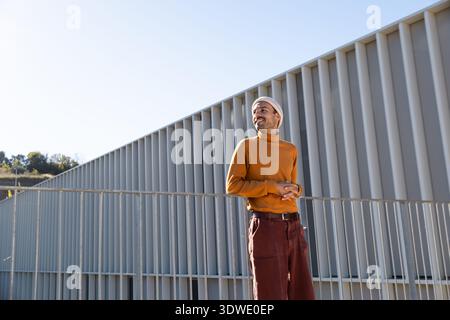 A man in an orange sweater and beanie stands outdoors in front of a modern, geometric building, looking thoughtfully into the distance on a sunny day. Stock Photo