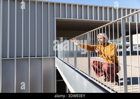 A man in an orange sweater and hat crouches by a metal railing, gazing through with a thoughtful expression. The outdoor setting features modern archi Stock Photo