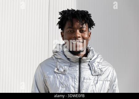 A confident man stands tall, wearing a sleek silver jacket that reflects a futuristic style. The minimal background and natural light highlight his ex Stock Photo