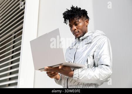 A man in a shiny silver jacket intently uses a laptop, examining its features against a modern backdrop, representing the integration of futuristic te Stock Photo