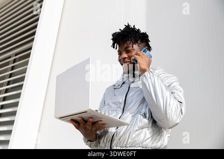 A man wearing a shiny silver jacket is standing outside, holding a laptop in one hand while talking on a smartphone. The setting appears modern and fu Stock Photo