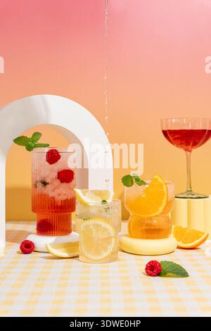 A vibrant display of cocktails featuring raspberries, oranges, and lemons, set against a pink and orange ombre background on a yellow checked tableclo Stock Photo