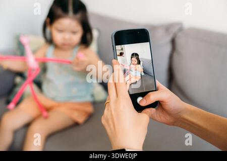 A person holds a smartphone, capturing a photo of a child playing with a pink toy on a couch. The child is out of focus, while the screen shows a clea Stock Photo