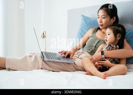 A mother and her child sit comfortably in bed while using a laptop. The child is holding a stuffed animal, creating a cozy and intimate atmosphere. Stock Photo