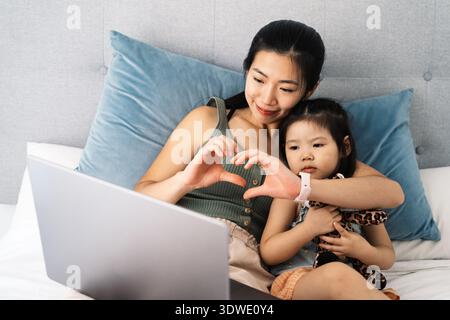 A mother and her child are sitting comfortably on a bed, engaged in watching a video on a laptop. The mother forms a heart with her hands while holdin Stock Photo