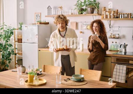 A redhead woman smiles while holding a warm drink, and her blonde partner serves croissants. Stock Photo