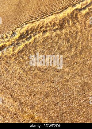 A close-up view of golden sand with gentle ripples formed by the clear water on the shore, capturing the tranquil beauty of a peaceful beach setting i Stock Photo
