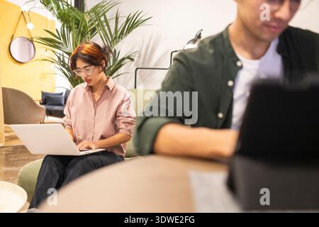 Modern coworking environment featuring two focused team members working on laptops. The setting includes comfortable seating, lush greenery, and a cal Stock Photo