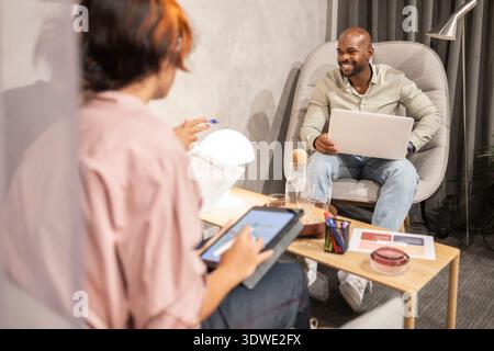 A team engaged in a productive coworking session. One person, smiling, holds a laptop, while another writes on a tablet. The modern office setup enhan Stock Photo