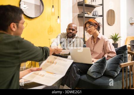 A diverse team collaborates in a modern coworking space. They engage in discussion and share creative ideas while working on laptops and reviewing doc Stock Photo
