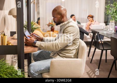 A diverse team working collaboratively in a modern coworking space. One person is focused on a tablet, while others engage in a lively discussion in t Stock Photo