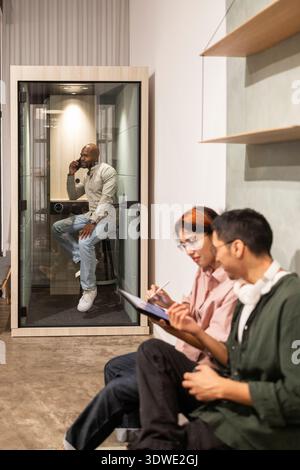 A modern coworking office featuring employees collaborating. One person is in a phone booth, while two others discuss and take notes, showcasing a ble Stock Photo