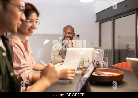 A diverse group of professionals collaborating in a modern coworking space. They are engaged in discussion around a table with laptops and documents, Stock Photo