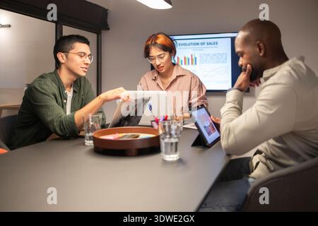Diverse team collaborating in a modern coworking office, engaging in a dynamic discussion with laptops and tablets, surrounded by charts and graphs on Stock Photo