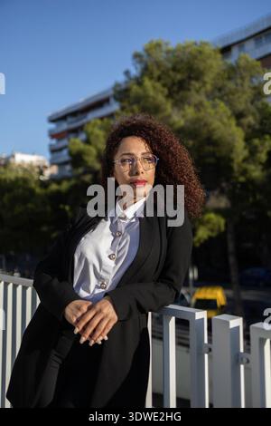 A confident business woman with curly hair stands outside, exuding professionalism. She wears glasses and is dressed in a smart business suit, leaning Stock Photo