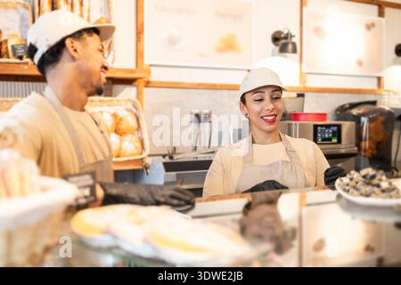 Two cafe workers share a light moment in a bustling bakery. Dressed in uniforms and aprons, they engage over pastries and bread, reflecting a friendly Stock Photo