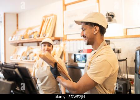 Two bakery workers in uniform happily serve fresh baked goods in a cozy cafe setting. Their teamwork and friendly demeanor create a warm and inviting Stock Photo