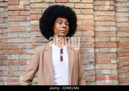 A stylish person stands confidently against a rustic brick wall, wearing a brown blazer over a white shirt, with sunglasses hanging from their shirt c Stock Photo