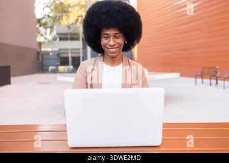 A person sits on an outdoor bench, smiling while using a laptop, dressed in a casual brown blazer. Bright sunlight and urban surroundings create a rel Stock Photo
