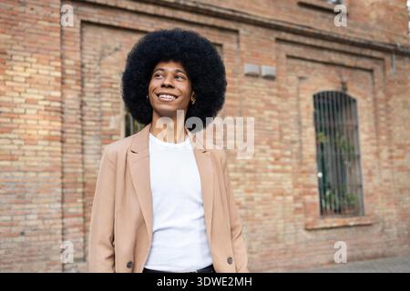 A person with an afro hairstyle is seen smiling while wearing a brown blazer and a white shirt. The background features a rustic brick wall, giving a Stock Photo