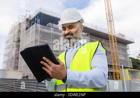 An engineer in a safety vest and hard hat examines construction plans using a tablet at a construction site. A building and crane are visible in the b Stock Photo