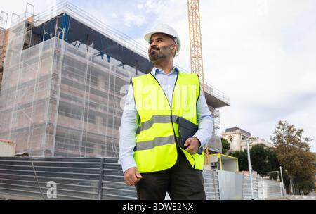 A confident construction engineer in a high-visibility vest and hard hat surveys a building site, overseeing the progress and ensuring safety standard Stock Photo
