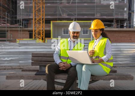 Two construction engineers in high-visibility vests and hard hats are intently discussing a project while looking at a laptop at a dynamic constructio Stock Photo