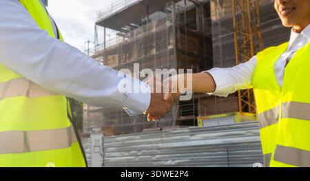 Two engineers wearing high-visibility vests shake hands at a construction site, symbolizing teamwork and collaboration. The building process continues Stock Photo