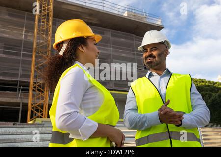 Two construction engineers in safety vests and helmets communicate on a job site, discussing project details. The background shows scaffolding and con Stock Photo