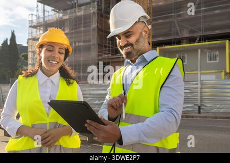 Two construction engineers wearing safety vests and helmets review project plans on a tablet. They stand in front of a building under construction, co Stock Photo