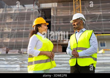 Two construction engineers in safety vests and helmets discuss project details on-site. The background shows scaffolding and a partially completed bui Stock Photo