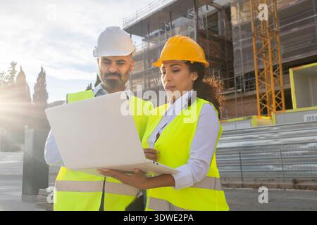 Two engineers wearing hard hats and reflective vests examine project details on a laptop at a construction site. A crane and scaffolding are visible i Stock Photo