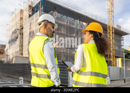 Two engineers wearing safety vests and helmets discuss a project at a construction site with building framework in the background while checking a dig Stock Photo