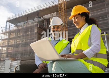 Two engineers in safety helmets and vests review construction plans on a laptop at a building site. They collaborate closely, ensuring project accurac Stock Photo