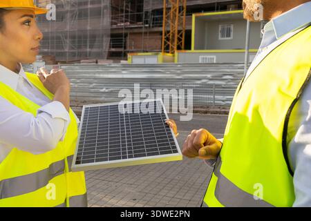 Two construction engineers in yellow safety vests and helmets examine a solar panel on site. They are discussing sustainable energy solutions for the Stock Photo