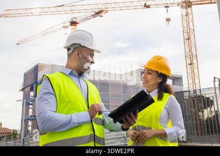 Two engineers wearing safety vests and helmets discuss construction plans on a busy site. They are surrounded by cranes and scaffolding, working colla Stock Photo